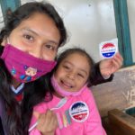 A woman holding a voting sticker, with her daughter a future voter.