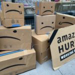 Amazon boxes with a sad arrow face are stacked on each other and a sign reading "Amazon hurts working people."